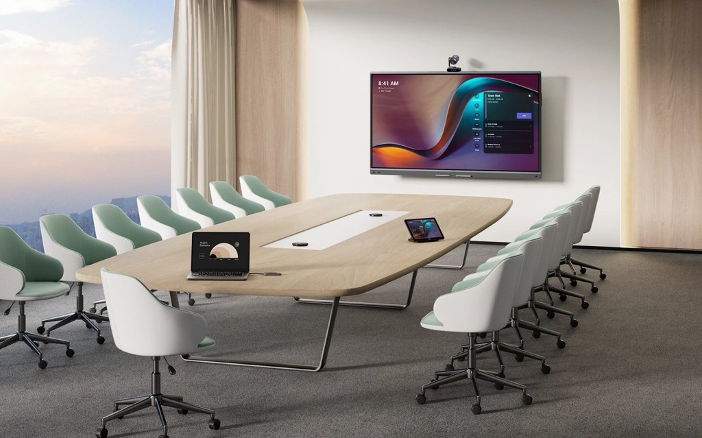 Sala de videoconferencias grande con cámara sobre la pantalla, panel táctil en la mesa y sillas para doce personas